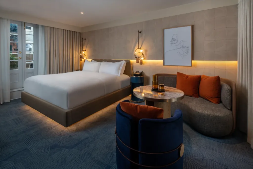 A modern hotel room featuring a large bed, round marble table, blue and gray chairs, wall art, soft lighting, and a window with curtains on the left.